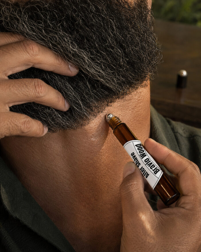 Mão aplicando after shave no pescoço de homem barbudo com frasco cilíndrico de vidro âmbar e esfera metálica deslizante com rótulo branco