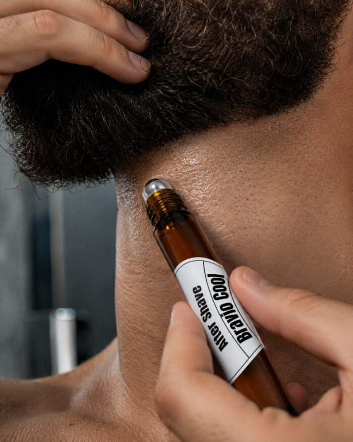 mão direita aplicando pós barba com frasco cilíndrico de vidro âmbar com ponta de espera metálica deslizante no pescoço em homem de barba