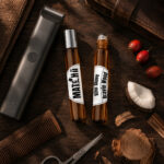 Dois frascos cilíndricos na vertical de vidro âmbar com after shave , um fechado com tampa prateada e outro com esfera metálica deslizante, em destaque rodeados por tesoura de alumínio pedaço de madeira de cedro e coco máquina de aparar barba e pente de madeira