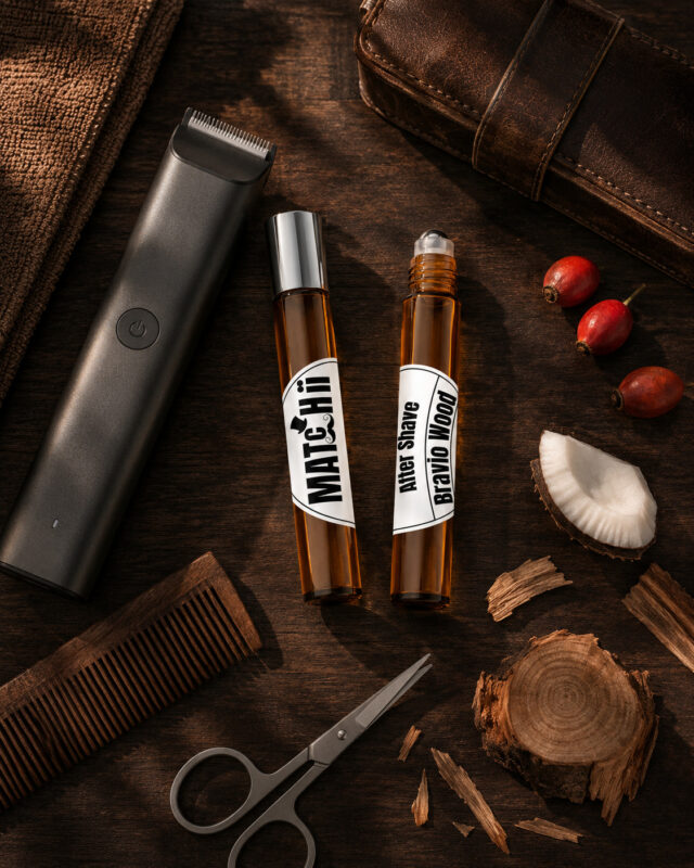 Dois frascos cilíndricos na vertical de vidro âmbar com after shave , um fechado com tampa prateada e outro com esfera metálica deslizante, em destaque rodeados por tesoura de alumínio pedaço de madeira de cedro e coco máquina de aparar barba e pente de madeira