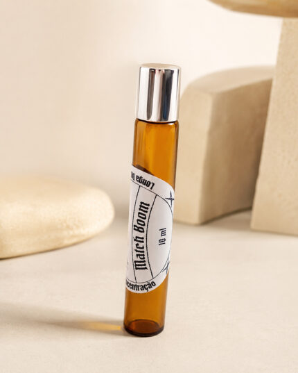 frasco de perfume roll on de vidro âmbar com tampa cromada na posição vertical com rótulo redondo branco match boom 10 ml em cima de tampo de granito bege ao fundo pedaços de pedra de granito