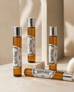 quatro frascos cilíndricos de perfume portátil feitos com vidro âmbar rótulos redondos brancos e tampas prateadas três na posição vertical e um na horizontal sob pedra de granito clara