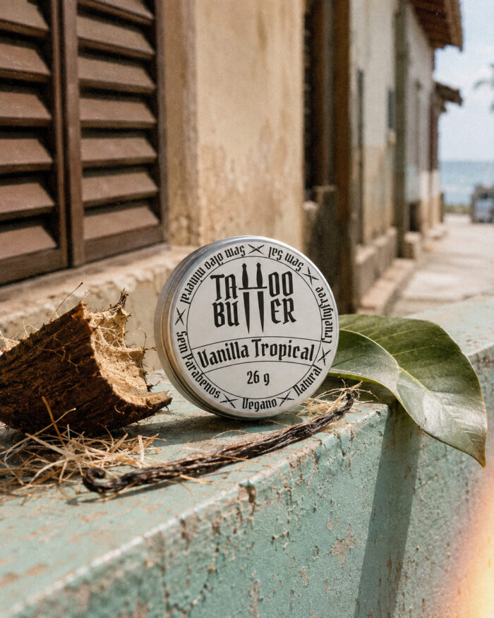 Latinha de alumínio Tattoo Butter Vanilla Tropical com rótulo branco redondo, apoiada sobre superfície desgastada, cercada por coco aberto, folha verde e fava de baunilha em cenário externo urbano.
