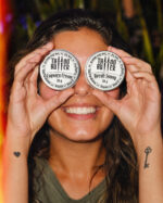 Mulher sorridente segura duas latinhas de alumínio Tattoo Butter diante dos olhos, com rótulos brancos redondos escritos Cupuaçu Cream e Neroli Sunny, em ambiente externo com vegetação ao fundo.