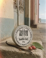 Latinha de alumínio Tattoo Butter Vanilla Soft com rótulo branco redondo em destaque, posicionada sobre muro de concreto, acompanhada por fava de baunilha, folha verde e pequenos frutos secos.
