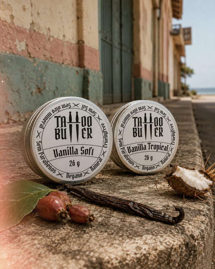 Duas latinhas de alumínio Tattoo Butter com rótulos brancos redondos escritos Vanilla Soft e Vanilla Tropical, lado a lado sobre muro rústico, com coco, baunilha, folha e frutos em primeiro plano.