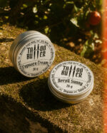 Duas latinhas de alumínio Tattoo Butter com rótulos brancos redondos Cupuaçu Cream e Neroli Sunny, apoiadas sobre uma superfície coberta de musgo sob luz quente natural.