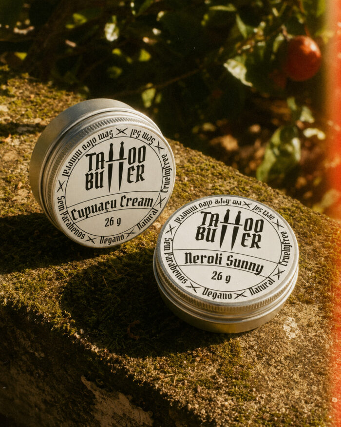 Duas latinhas de alumínio Tattoo Butter com rótulos brancos redondos Cupuaçu Cream e Neroli Sunny, apoiadas sobre uma superfície coberta de musgo sob luz quente natural.