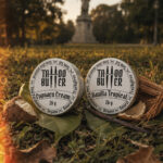 Duas latinhas de alumínio do Kit Ikii, com rótulos brancos redondos escritos Tattoo Butter Cupuaçu Cream e Vanilla Tropical, apoiadas sobre folhas e grama em um parque, com coco ao redor e estátua desfocada ao fundo.