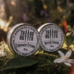 Duas latinhas de alumínio Tattoo Butter com rótulos brancos redondos escritos Cupuaçu Cream e Neroli Sunny, posicionadas sobre musgo em ambiente de mata com flores brancas e folhas ao redor.