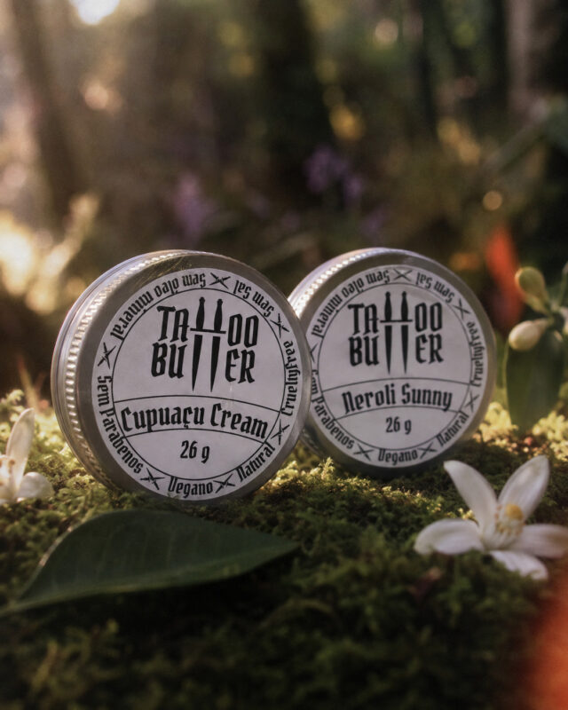 Duas latinhas de alumínio Tattoo Butter com rótulos brancos redondos escritos Cupuaçu Cream e Neroli Sunny, posicionadas sobre musgo em ambiente de mata com flores brancas e folhas ao redor.