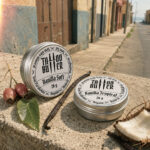 Duas latinhas de alumínio Tattoo Butter, com rótulos brancos redondos escritos Vanilla Soft e Vanilla Tropical, apoiadas sobre muro de concreto com baunilha, coco, folha e frutos ao redor.
