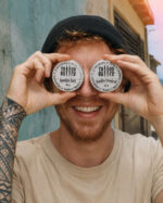 Homem sorridente segura duas latinhas de alumínio Tattoo Butter diante dos olhos, com rótulos brancos redondos Vanilla Soft e Vanilla Tropical, em cena externa com rua ao fundo.