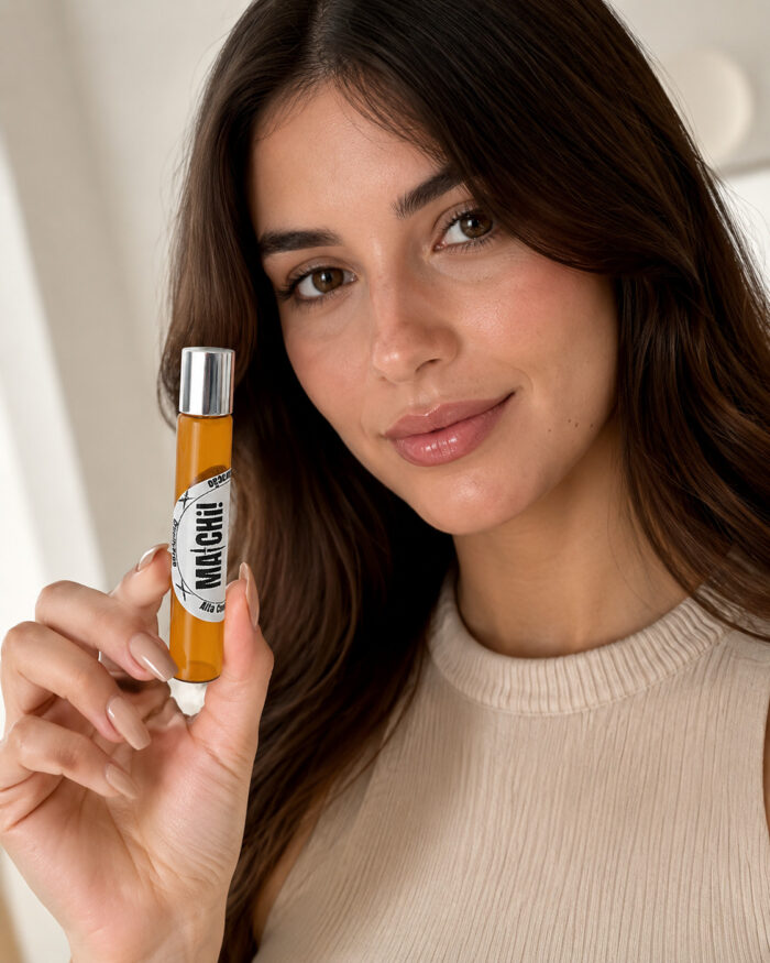 mulher branca feliz cabelo castanho contato visual direto segurando com mão direita ao lado do rosto frasco de perfume de bolsa feito com vidro âmbar com tampa cromada rótulo branco matchii