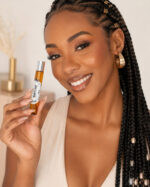 modelo negra com tranças sorrindo vestindo roupa bege contato visual direto segurando frasco de perfume feminino em formato cilíndrico de vidro âmbar com tampa cromada perto do rosto
