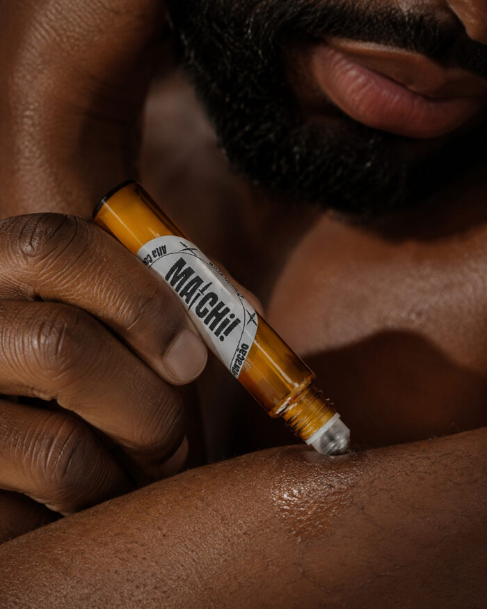 parte do rosto de homem com barba e mão aplicando perfume no antebraço com frasco cilíndrico feito com vidro âmbar com ponta de esfera redonda metálica deslizante