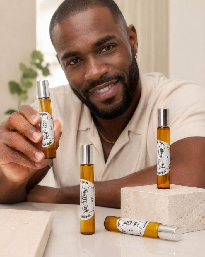 homem negro com barba sorrindo contato visual direto segurando frasco cilíndrico de perfume portátil feito com vidro âmbar tampa prateada e rótulo branco redondo e mais dois frascos na vertical e um na horizontal sob tampo de granito claro