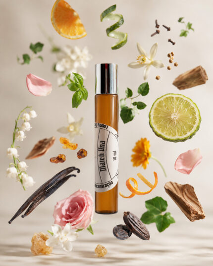 perfume feminino no frasco cilíndrico em vidro âmbar com tampa cromada com rótulo branco redondo match una com fatia de limão rosa fatia de laranja fava tonka lírio cravo ao redor do frasco